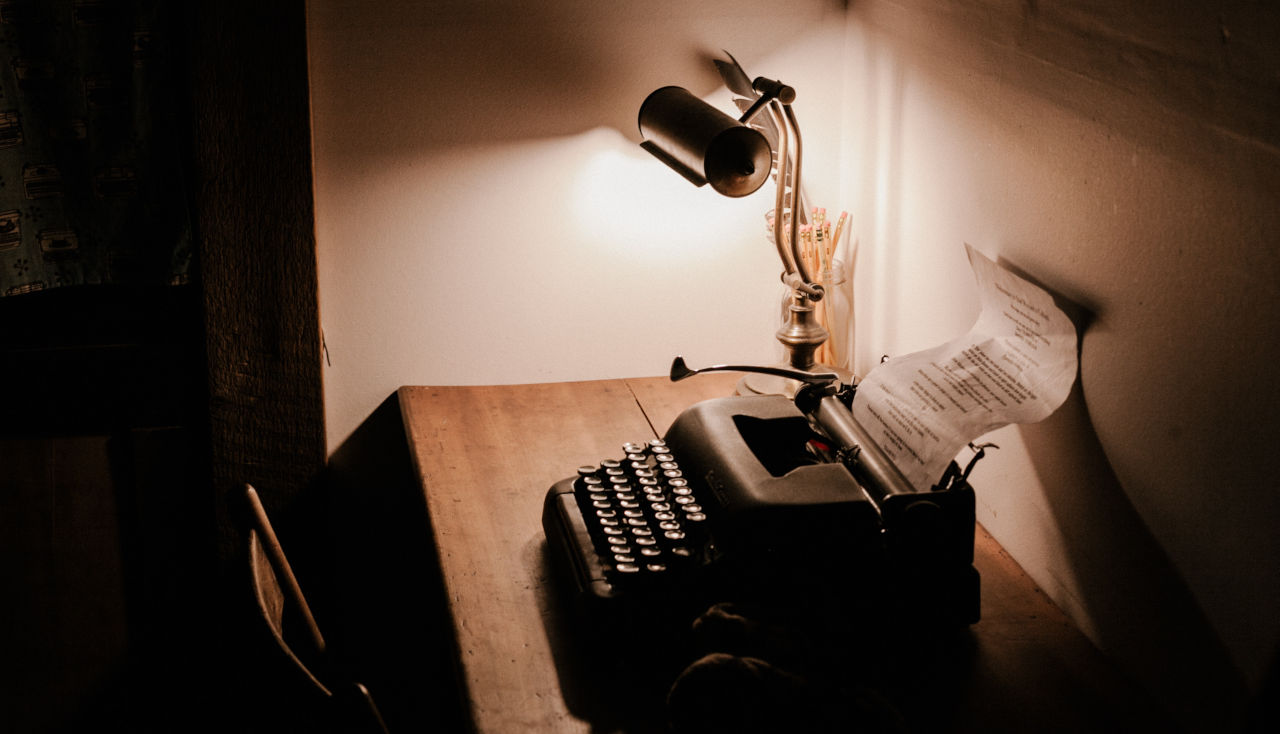 A desk in a dark room with a desk-lamp illuminating the area. A typewriter sits next to it.