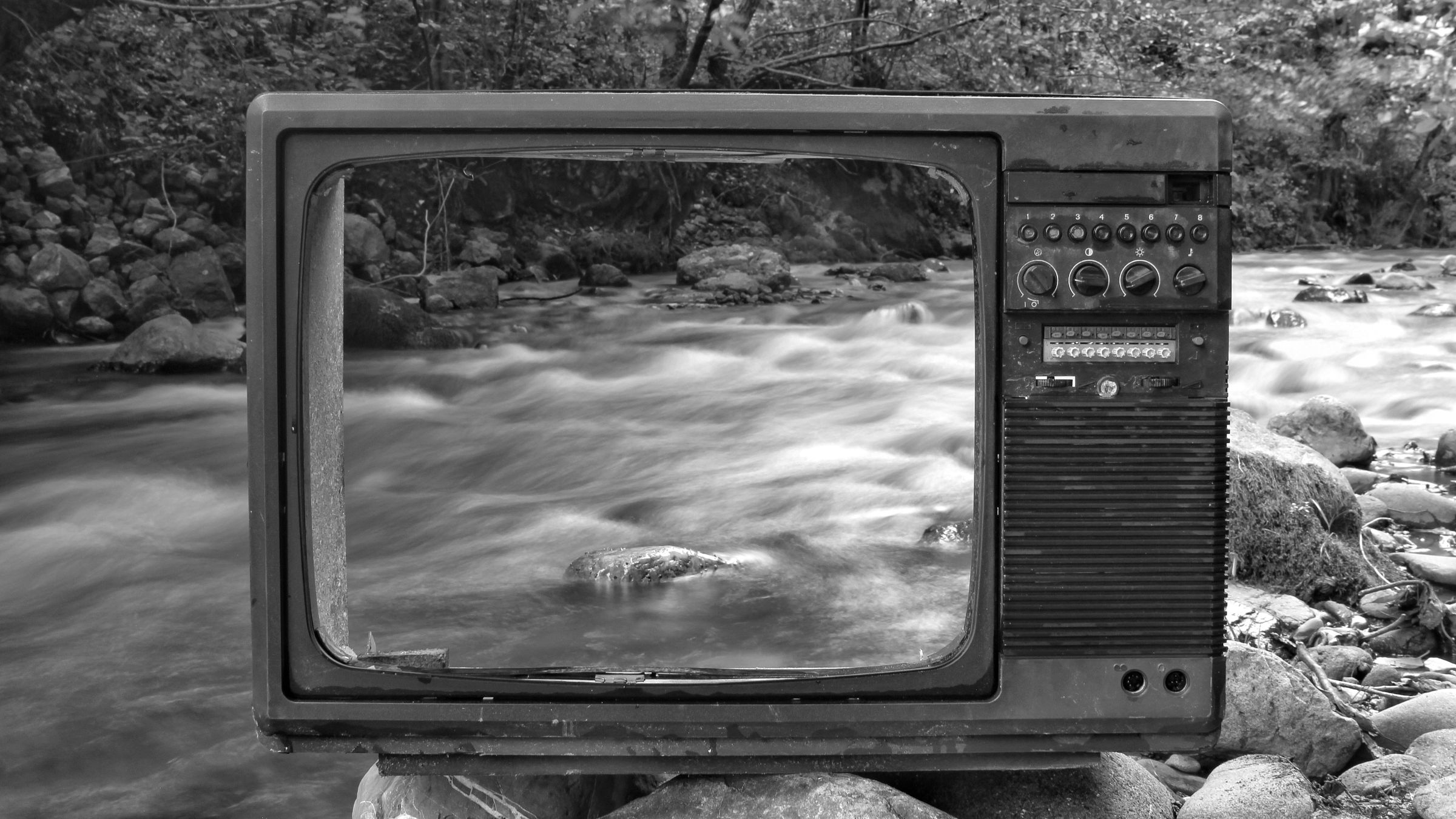 Television with a broken screen sitting on some rocks in a stream.