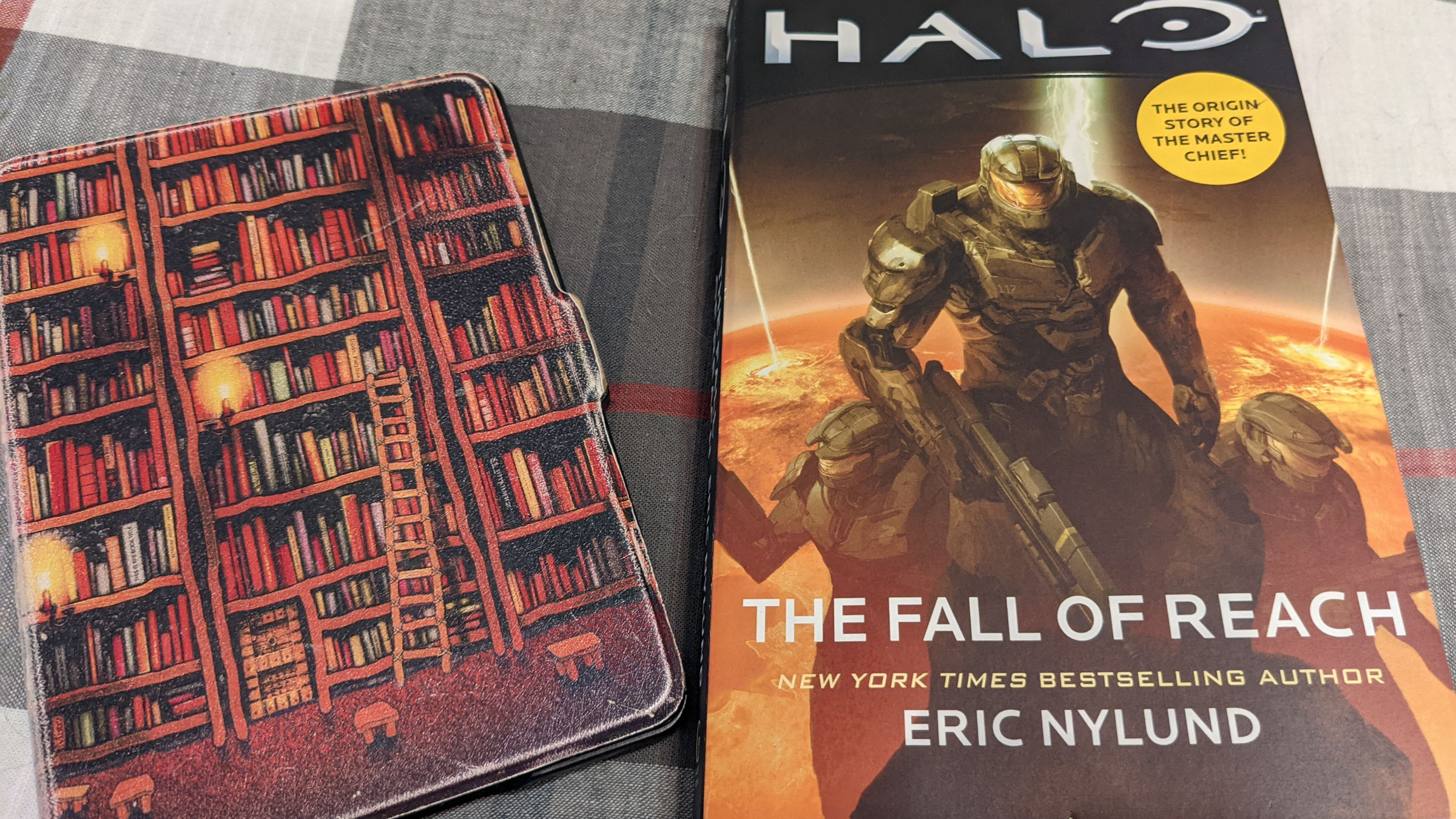 Amazon Kindle and paperback copy of ‘Halo: The Fall of Reach’ lying on a plaid bedspread.