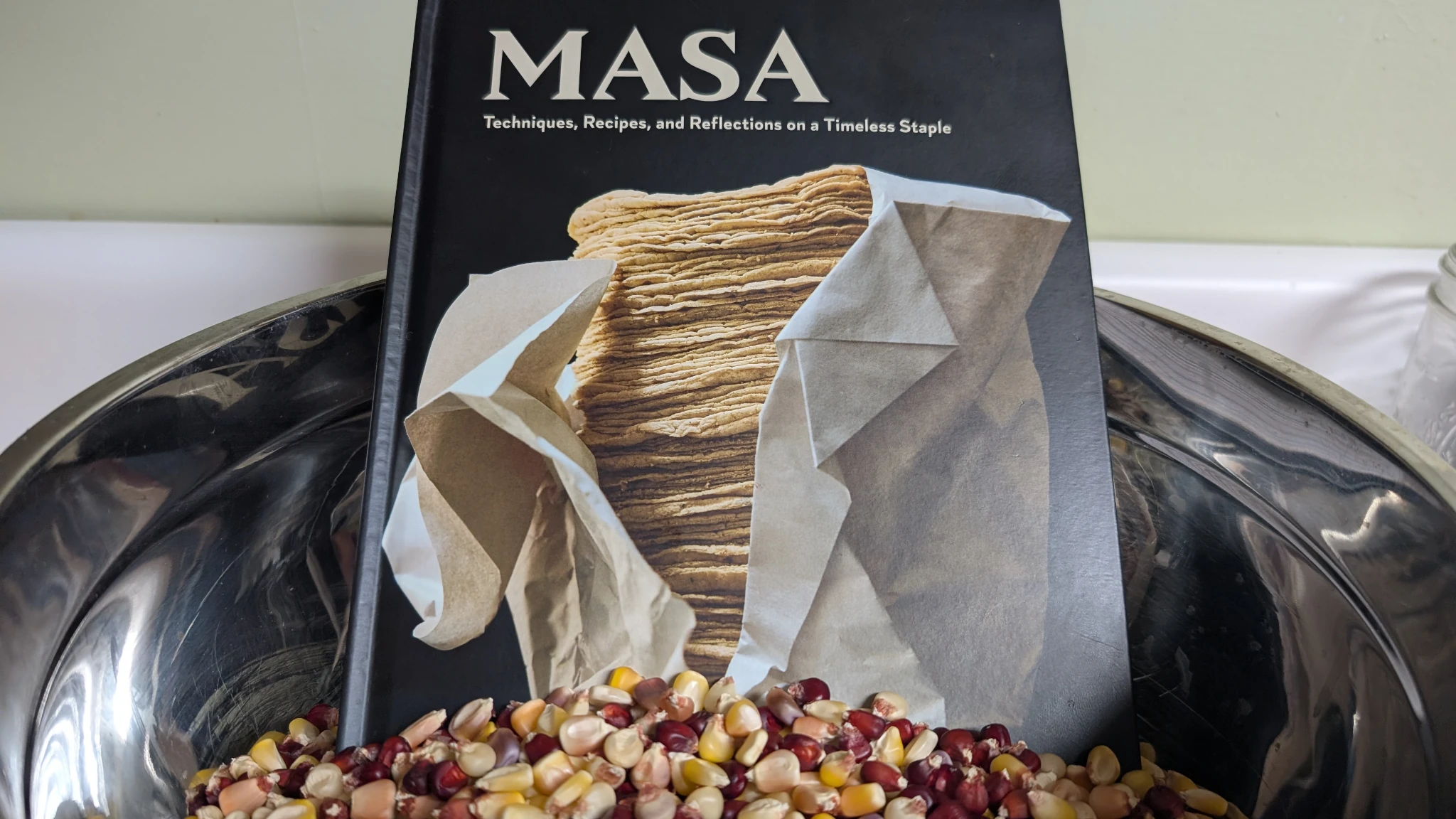 A metal bowl of colorful dent corn, yellow and red, with a book titled Masa sitting on top.